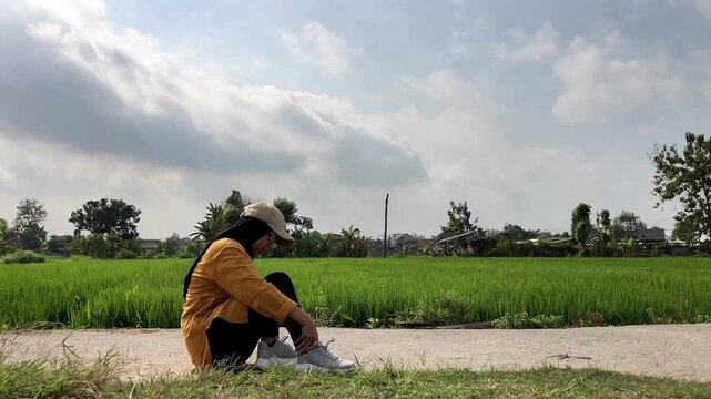 Slow motion clip of a Muslim woman in a hijab sitting on a path by a green rice field. She is holding her foot and wincing in pain after experiencing an ankle sprain while jogging outdoors.