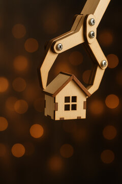 Wooden robotic claw gripping miniature house model with warm bokeh lights background, symbolizing futuristic property selection and real estate