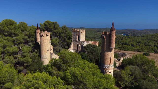 Aerial view of Castell de la Muga in Catalonia, Spain, featuring two conical-roofed medieval towers and a ruined central tower amid dense pine forest with coastal hills in the distance