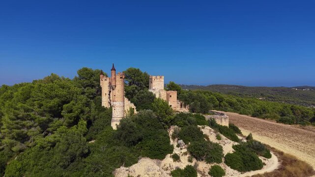 Aerial view of Castell de la Muga, a ruined medieval castle with conical-roofed towers rising above dense pine forest, dry farmland and hazy coastal hills in Catalonia, Spain