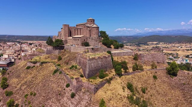 Aerial view of Cardona Castle atop a hill in Catalonia, Spain, featuring its Romanesque church, rounded apse, defensive star-shaped walls, and layered mountains in the distance
