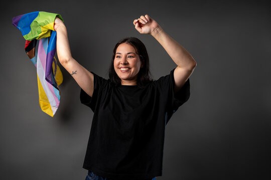 Non binary person on a grey background twirling a LGBTQA+ flag with their hands up, looking away from the camera with a smile.