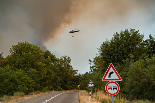 Firefighting Airbus H215 Super Puma helicopter flying with a Bambi bucket over a country road into dense wildfire smoke in Deliblatska Pescara. Deliblato, Serbia - August 7, 2026.