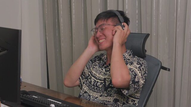 Young man laughs, adjusting headphones with joy. He sits at desk, wearing glasses and patterned shirt. Computer monitor visible, keyboard in front of him