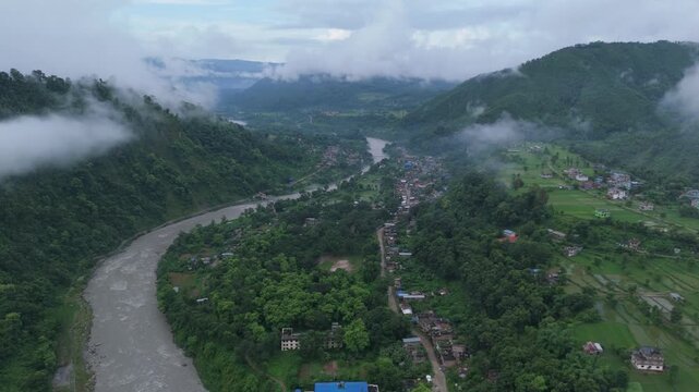 Sweeping drone view of a curving muddy river and scattered rural settlements nestled in a mist-covered green valley in Nepal