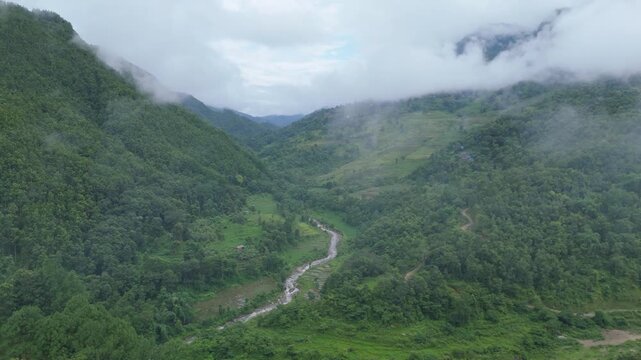 Wide drone establishing shot of a deep green Himalayan valley with a small river below and heavy morning mist clinging to the peaks in Nepal