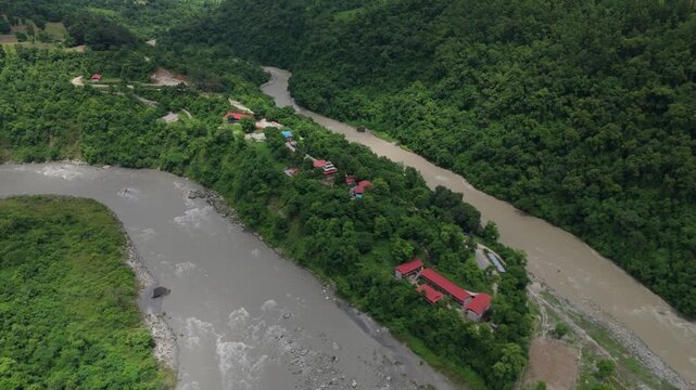 Aerial view of two rivers converging beside dense green forest and rural buildings in a mountainous landscape