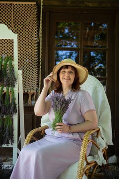  Woman sitting in a wicker chair on a cottage porch, holding a lavender bouquet while wearing a sunhat. She smiles warmly, surrounded by hanging lavender bunches and rustic decor