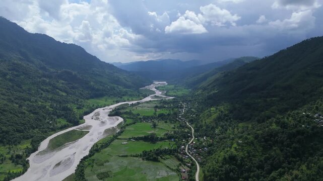 Aerial view of a braided muddy river cutting through a deep green mountain valley alongside a rural road
