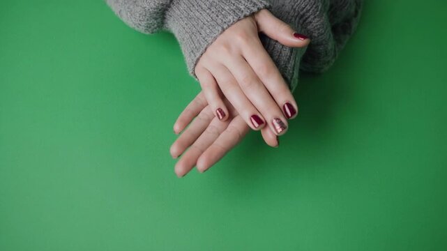Woman's hands with dark nail polish gently interlocking fingers on a bright green background with soft lighting