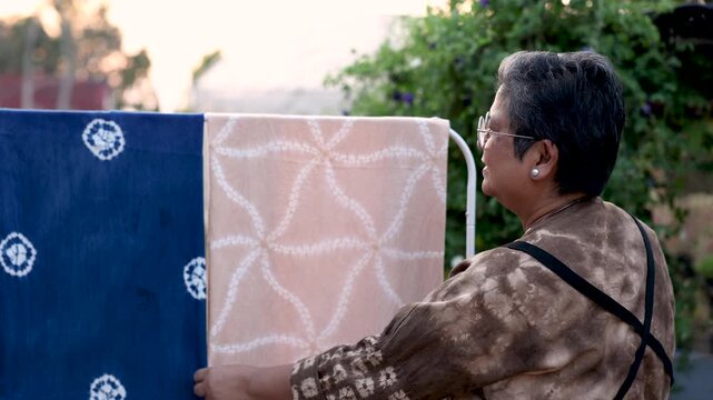 Healthy senior woman arranging colorful dyed fabrics on drying rack outdoors, textile craft business, creative skill, active retirement, productive aging, independent work lifestyle.