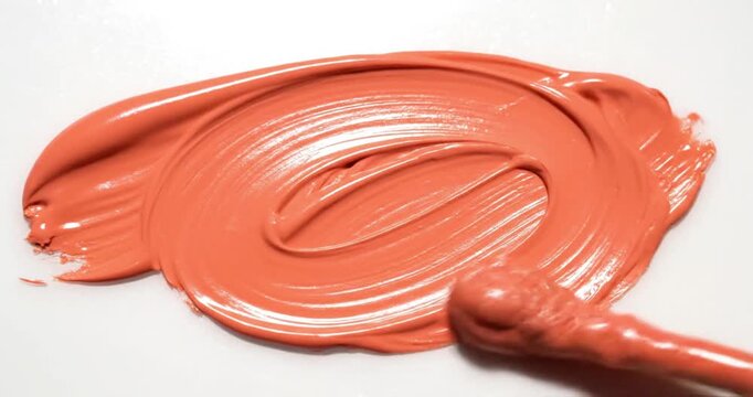 Coral-colored cosmetic cream being smeared on a white surface, thick liquid texture application, beauty and skincare product swatch.