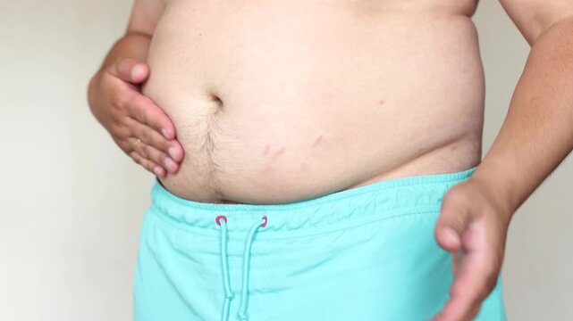 Slow motion video shows man tapping large belly with palms while wearing blue shorts. Footage explores body weight challenges.