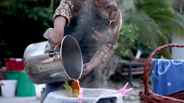 Senior artisan entrepreneur pouring hot natural dye into filtered container outdoors, textile production, craft business workflow, active retirement, productive aging lifestyle business.