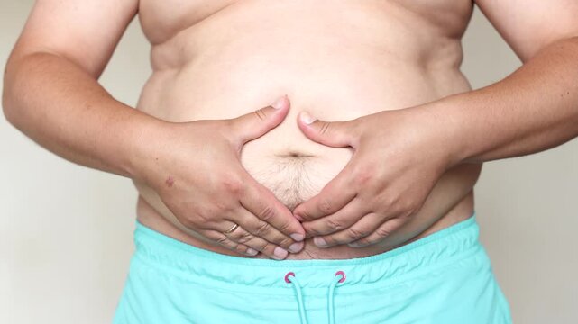 Slow motion of young man at medical checkup touching his large belly. Scene suggests excess belly fat linked to high sugar intake and frequent sugary soda consumption.