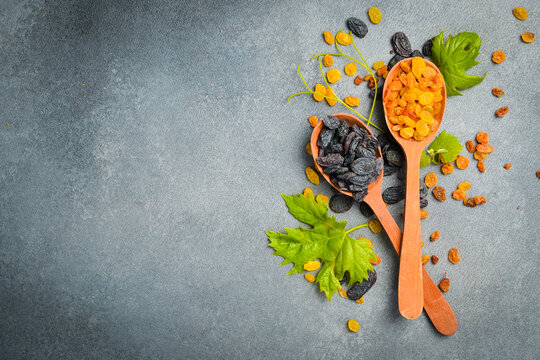 Golden and dark dried raisins fill wooden spoons on a grey slate background. Fresh green grape leaves garnish sweet fruits. Healthy organic food concept.