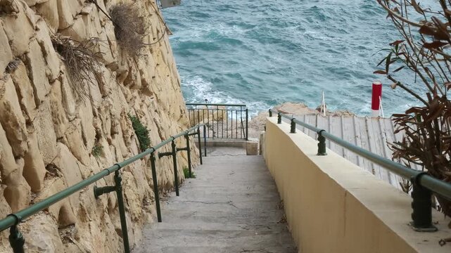 stone staircase descends cliffside toward open sea railings guiding path overcast valletta coastline waves visible below rugged limestone edge malta coastline mediterranean sea sea spray