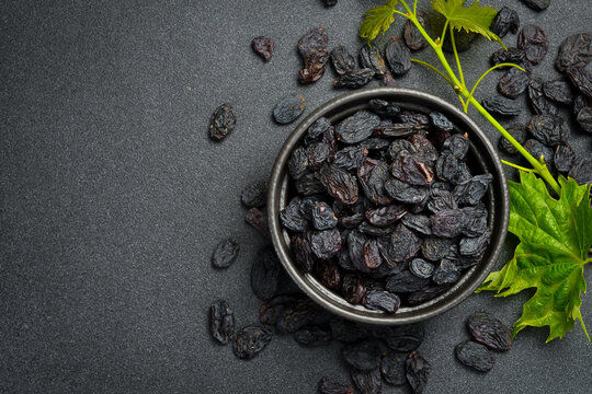 Top view of dark raisins inside a rustic bowl on grey stone surface. Delicious dried grapes are scattered around the bowl. Great image for food.