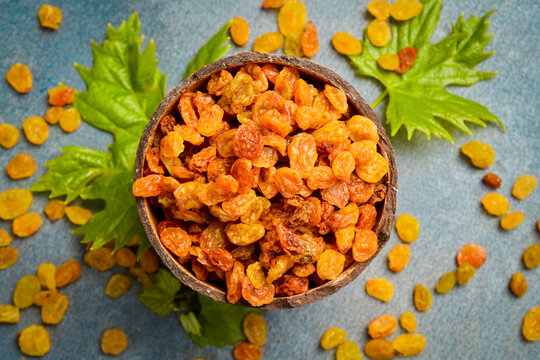 Golden dried raisins fill a round wooden bowl placed on a grey slate background. Fresh green grape leaves garnish the sweet delicious fruits. Healthy food.