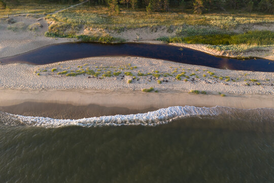Aerial view of a small river flowing across sandy beach into Baltic Sea at Perakula Nova in Estonia during warm morning golden hour light.