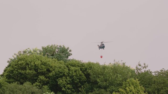 Tracking shot of a red Kamov Ka-32 firefighting helicopter flying towards camera with a Bambi bucket during wildfire operations in Deliblatska Pescara. Deliblato, Serbia - August 7, 2026.