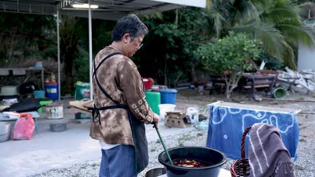Senior small business owner stirring dyed fabric outdoors, artisan textile production, hands on craft skill, self employment, active retirement, productive aging, creative lifestyle.