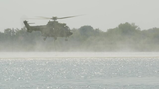 Medium close shot of an Airbus H215 Super Puma firefighting helicopter arriving to refill its Bambi bucket on a lake and flying away in Deliblatska Pescara. Deliblato, Serbia - August 7, 2026.