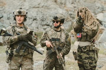 Fototapeta premium Outdoor military concept. Three focused soldier standing together feeling determined while wearing camouflage uniform and protective gear with rifle in rocky area
