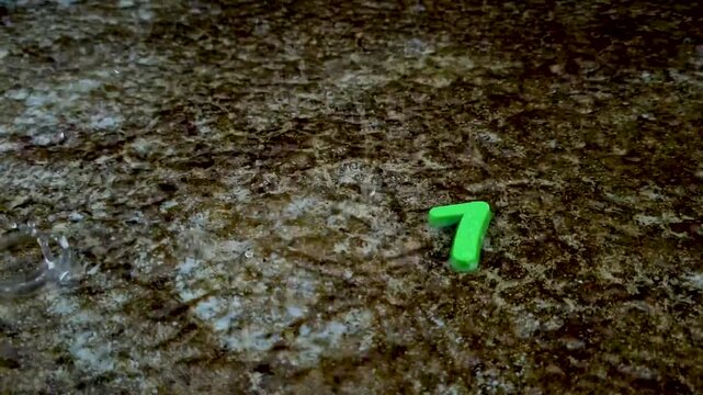 Green number seven toy on wet rough ground during heavy rain pavement water droplets storm texture