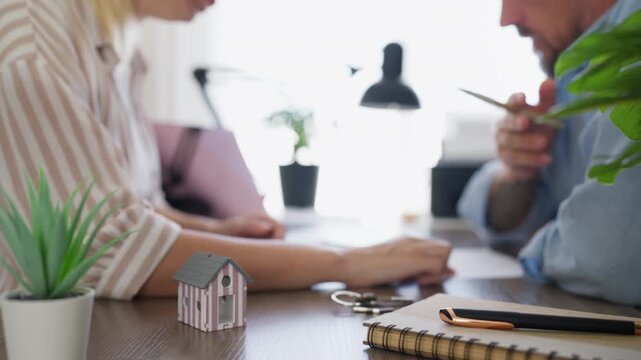 House model and keys in sharp focus on an office desk while a client reviews documents in the background. Home buying, property ownership, real estate services and purchase decision concept.