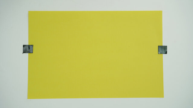 A piece of yellow paper positioned horizontal and attached to a surface with black tape on two sides, isolated on a white background.