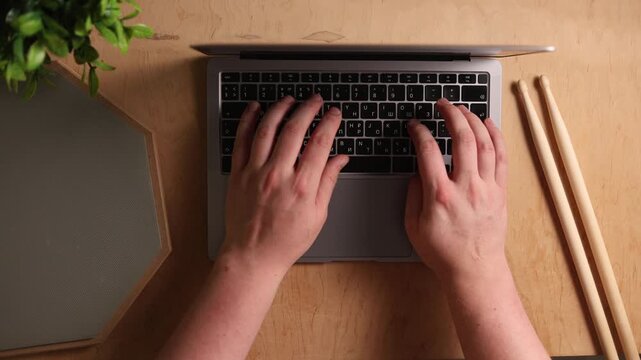 Overhead view of hands typing on a laptop keyboard next to drumsticks