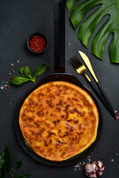 Traditional roasted round pie baked inside black frying pan top view. Tasty golden cheese flatbread surrounded by garlic and chili peppers. Gourmet homemade meal recipe.