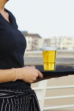 Server holds tray with golden beer. Bubbles rise gently in clear glass. Background hints at waterfront lounge. Neutral expression, focused on service