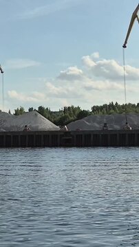 Industrial crane loading gravel at a river port. Cargo ship view of harbor docks with sand piles and construction machinery during summer trip on the water.