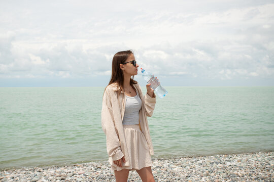 teenage girl drinks water on a pebble beach by the sea, promoting hydration, heat safety, healthy summer travel and refreshment during hot coastal weather
