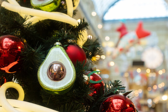 playful avocado christmas ornament hanging on evergreen branches among red baubles and fairy lights,modern quirky holiday decor in a bright indoor shopping arcade with bokeh