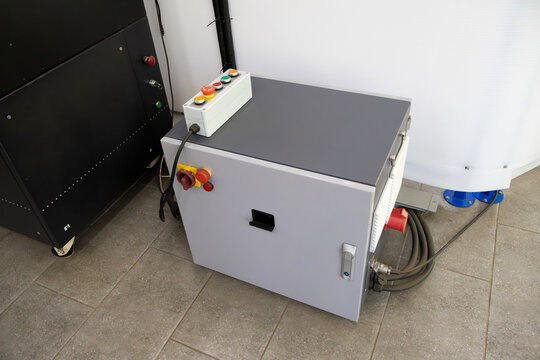Rugged metal cabinet hosts critical controls; white remote panel features red and yellow emergency buttons. Visible hoses and adjacent equipment convey active industrial environment