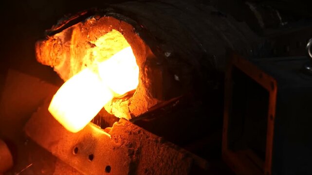Hot metal forging process featuring glowing glowing steel components being heated inside a heavy industrial furnace and shaped using automated mechanical equipment within a manufacturing plant setting