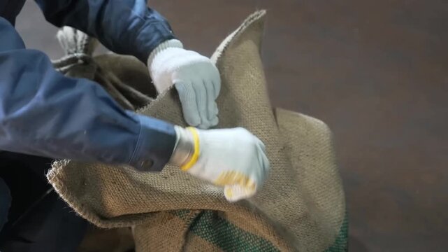 A factory worker wearing protective gloves uses pliers to twist and secure metal wire around the top opening of a heavy burlap sack in an industrial workshop environment