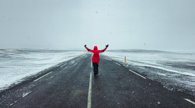 Lone Person in red jacket walking on a long, empty road during a snowstorm, holding arms wide, feeling freedom and adventure. Iceland stormy weather