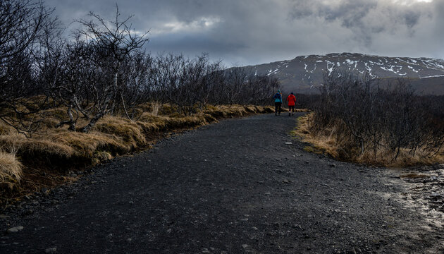 Two hikers walking gravel burrafoss path through a rugged landscape, with a snow capped mountain under a cloudy sky. Iceland Icelandic hiking trail