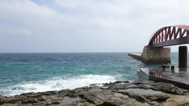 red arched breakwater bridge viewed from ground level waves surge against its stone pillar valletta harbour mouth valletta breakwater st elmo breakwater harbour entrance lighthouse tower
