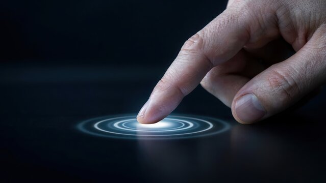 Close up of a human finger touching a glowing digital button on a dark surface creating circular light ripples representing touch screen technology