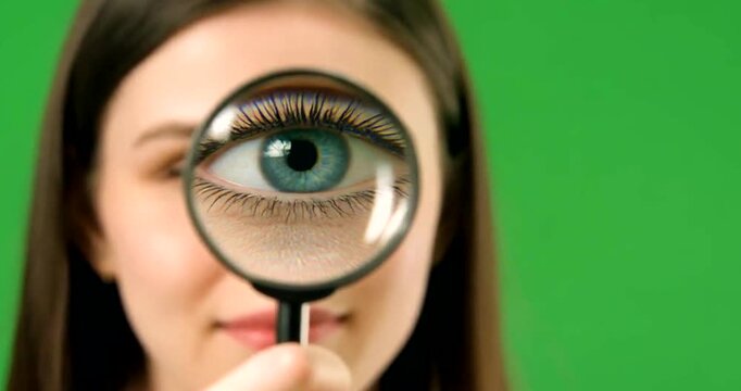 Woman using magnifying glass over eye against green screen for close inspection concept.