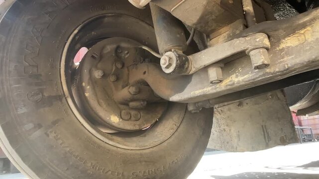 Close view of a dusty truck wheel hub, suspension linkage, and undercarriage components, showing bolts, metal joints, grease, and grime during an open-air heavy vehicle inspection carefully.