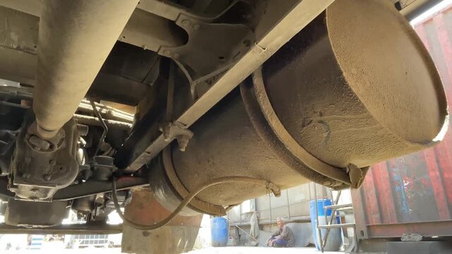 Close view of a truck exhaust system and cylindrical muffler beneath the chassis, showing dustier metal surfaces, pipes, brackets, and mechanical components during inspection at a workplace.