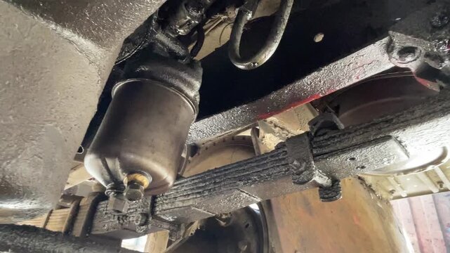 Detailed view beneath a truck showing the chassis, suspension, exhaust, and metal undercarriage components, with dirt and grease visible during an open-air mechanical inspection today, here.