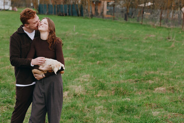 Obraz premium A couple in casual clothes share a kiss while holding a goat kid in a sunny meadow, capturing a warm rural lifestyle moment and affection in an open field.