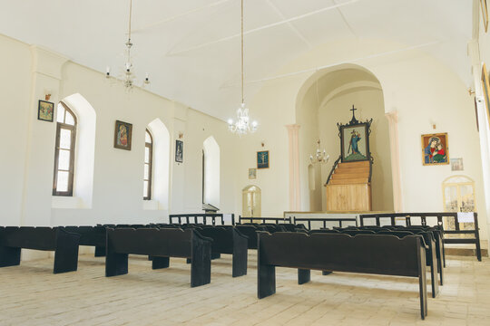 Interior view of Angeli Church in Hamedan, Iran, featuring rows of dark wooden pews facing an elevated altar with hanging chandeliers, arched windows, and religious artwork on white walls.